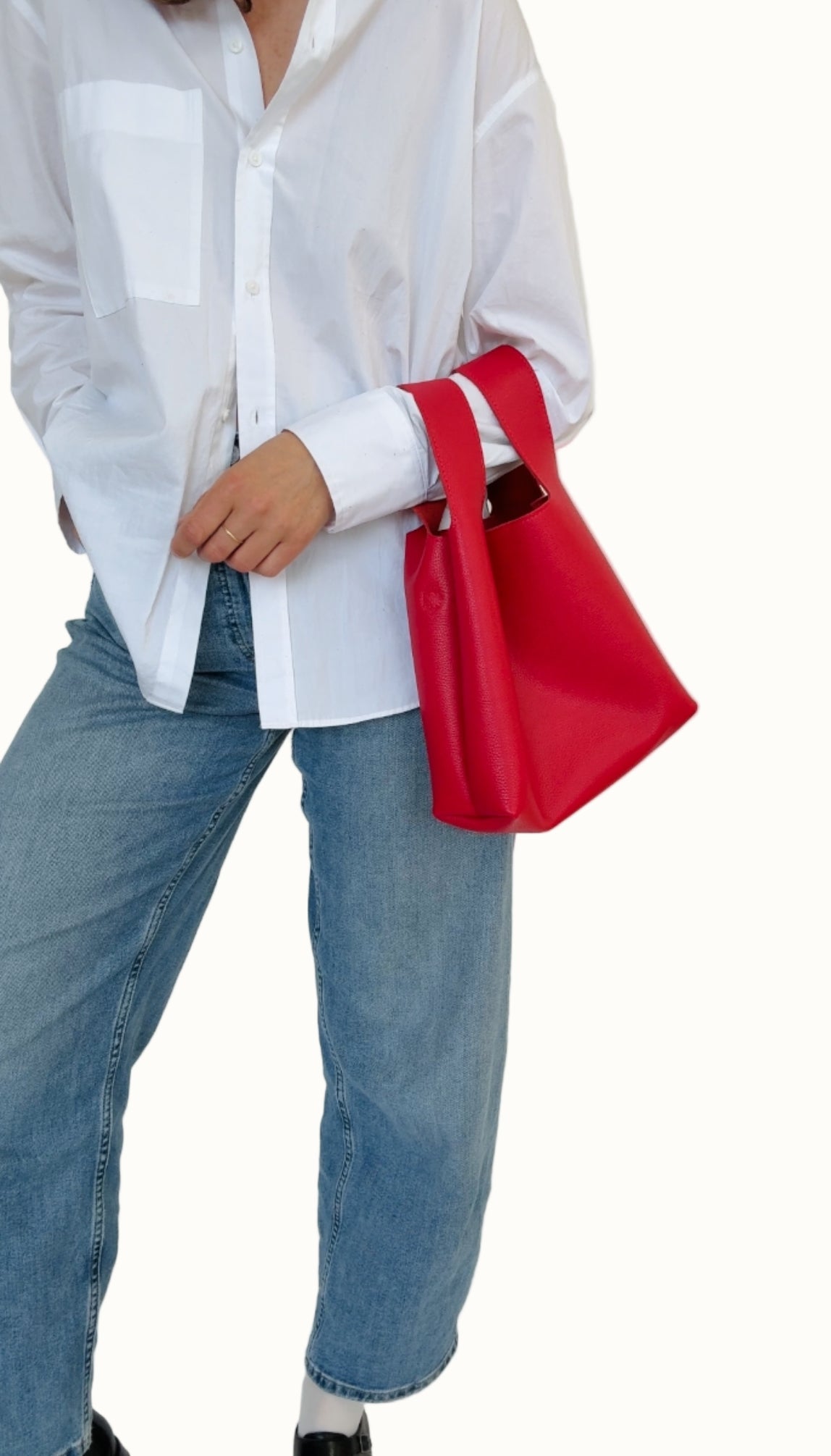 A person wearing a white shirt and blue jeans is holding a red mini tote bag with a shoulder strap.