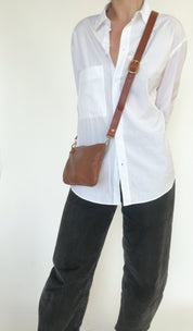 Person wearing a white shirt and black pants with a brown crossbody bag on a plain background