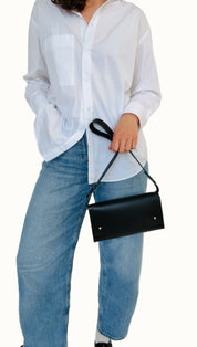 A person wearing a white shirt and blue jeans, holding a black shoulder bag with a strap.