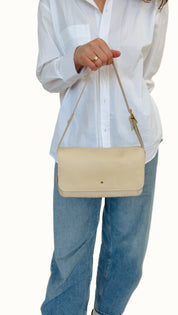 A person wearing a white shirt and blue jeans is holding a cream-colored crossbody bag with a shoulder strap and a zipper closure.