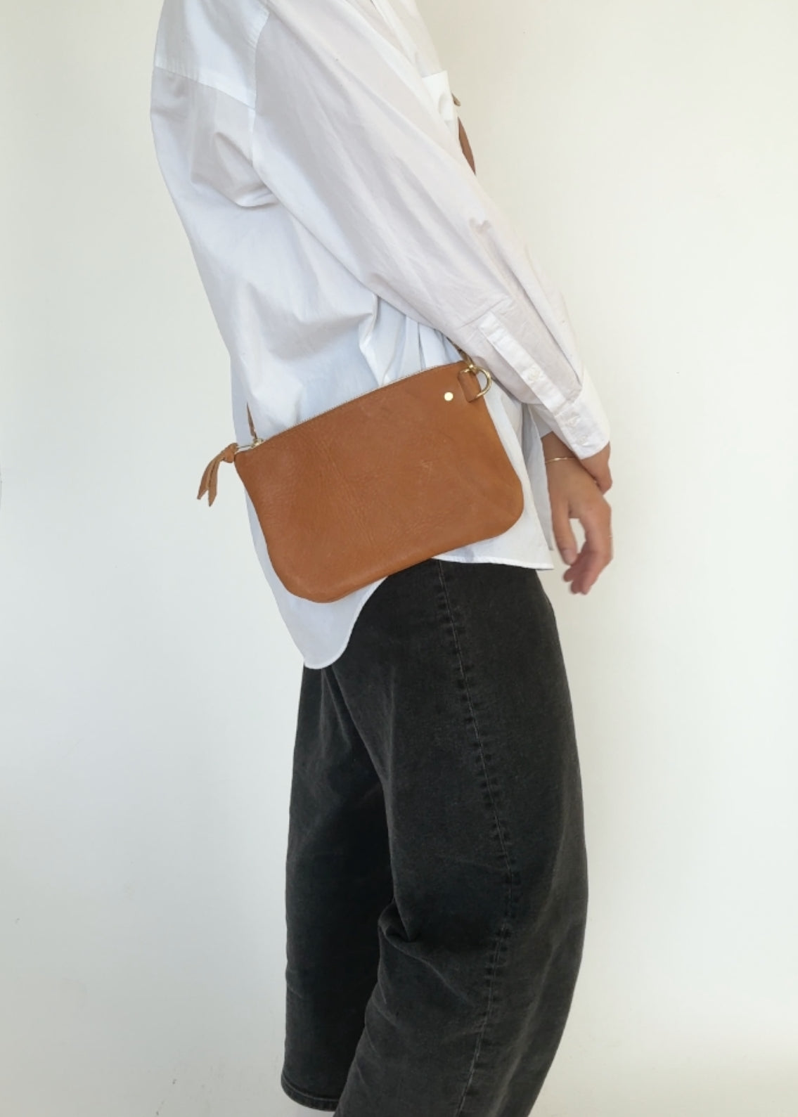 Person wearing a white shirt and black pants with a brown leather bag on a plain background