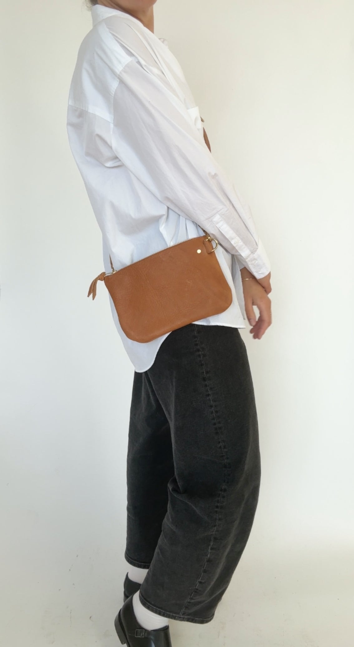 Person wearing a white shirt and black pants with a brown leather bag on a plain background