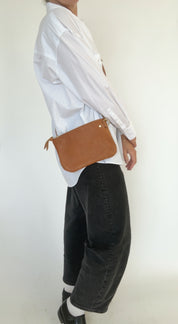Person wearing a white shirt and black pants with a brown leather bag on a plain background