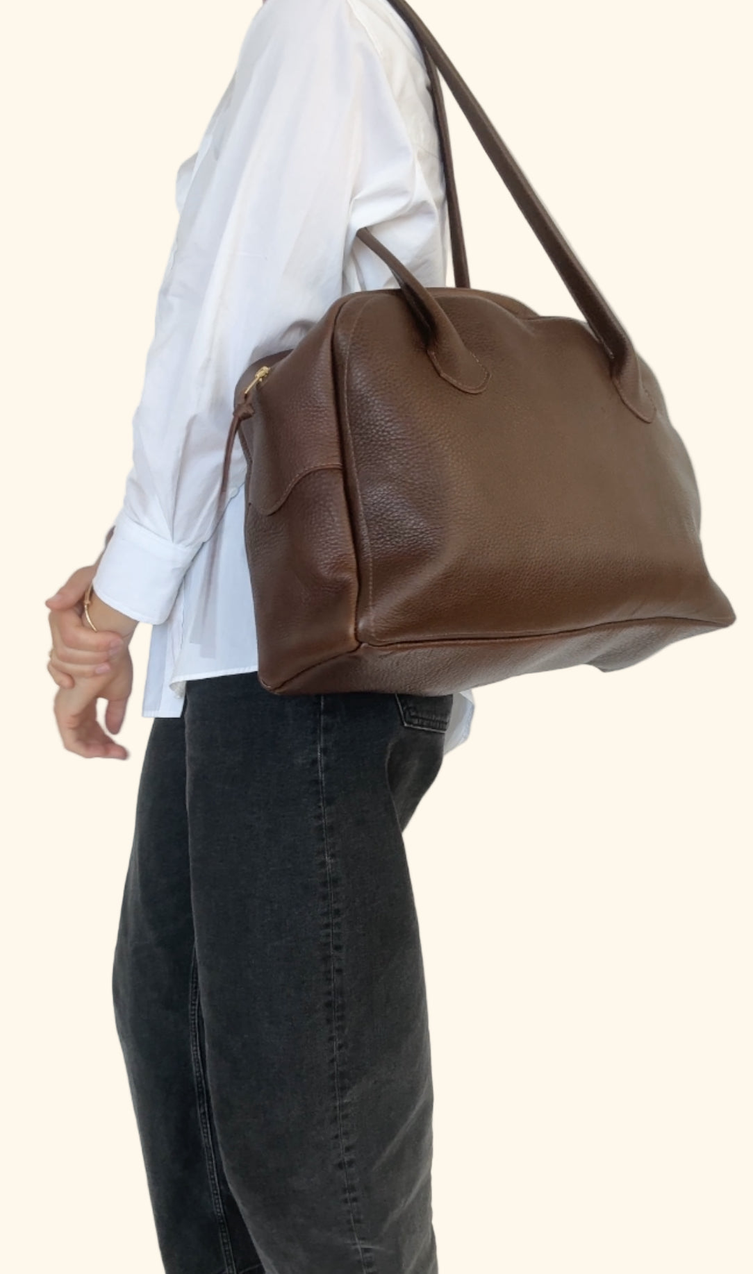 Brown leather handbag being held by a person wearing a white shirt and dark jeans on a plain background