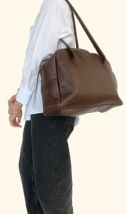 Brown leather handbag being held by a person wearing a white shirt and dark jeans on a plain background