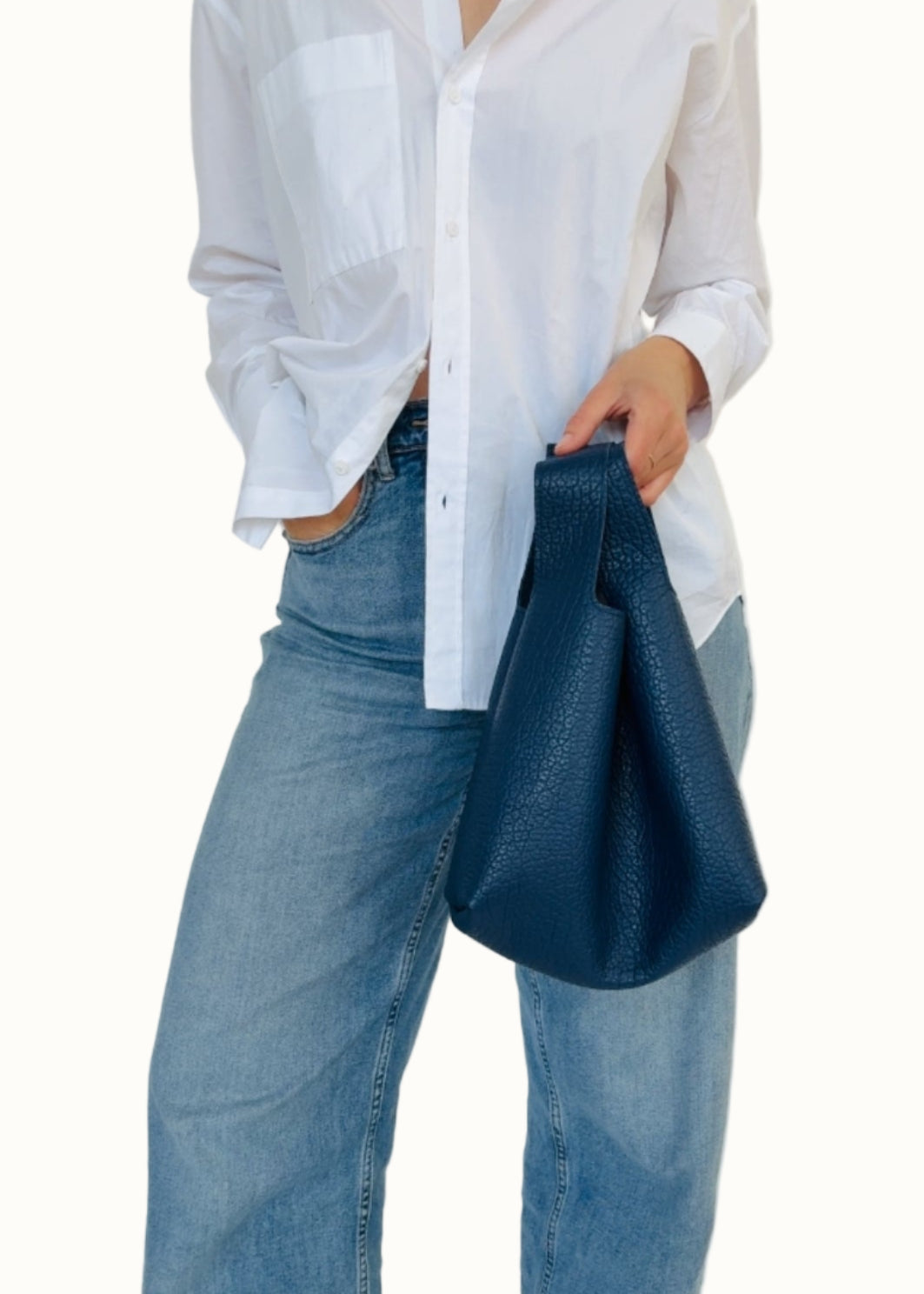 A person wearing a white shirt and blue jeans, holding a navy-colored mini tote bag with a leather shoulder strap.