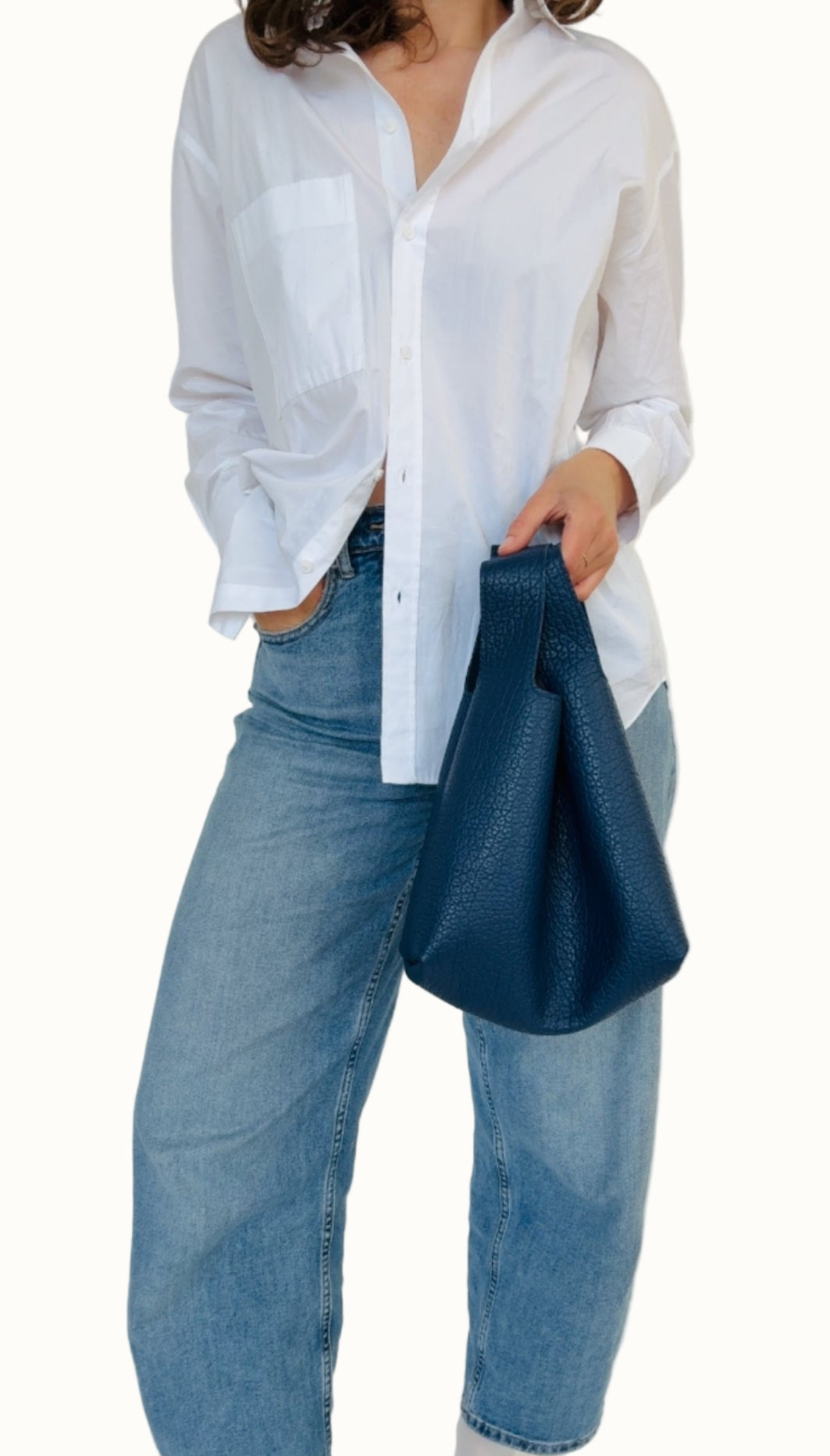 A person wearing a white shirt and blue jeans, holding a navy-colored mini tote bag with a leather shoulder strap.