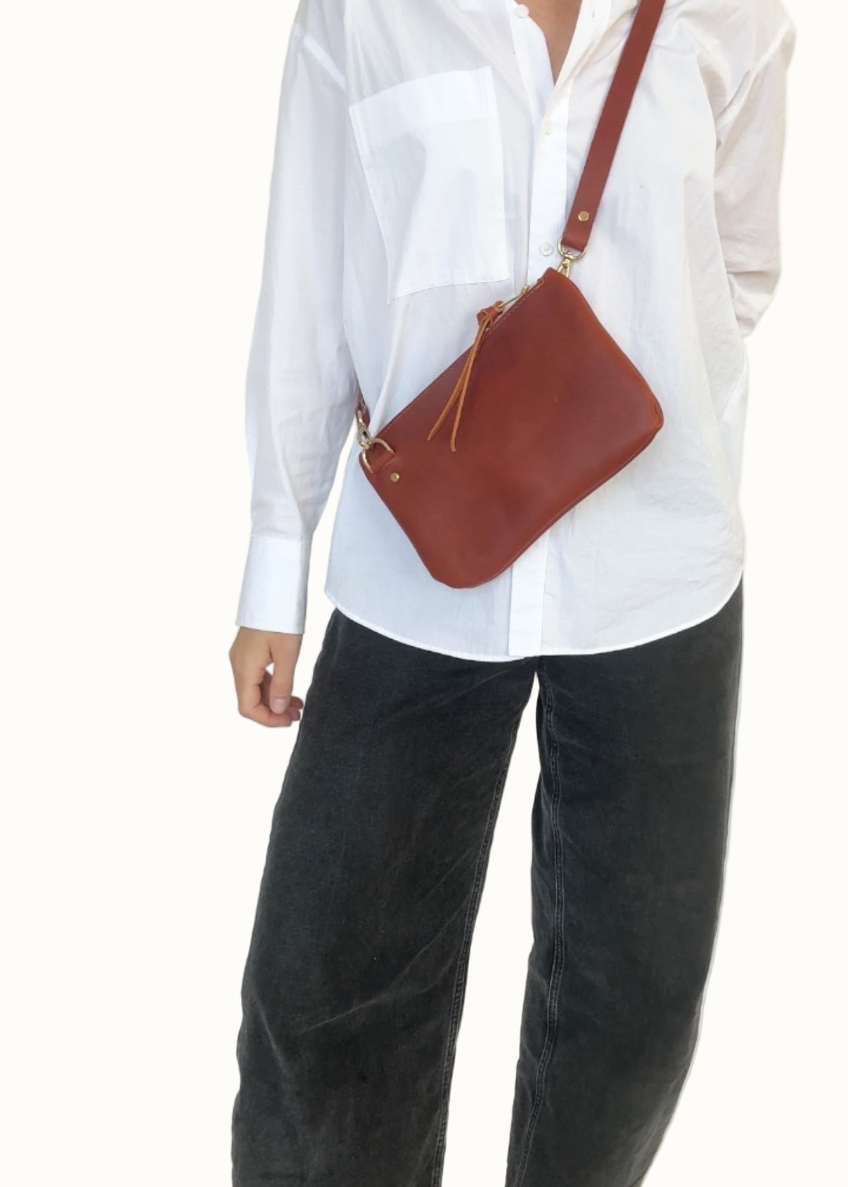 Person wearing a white shirt and dark pants with a brown leather bag on a plain background