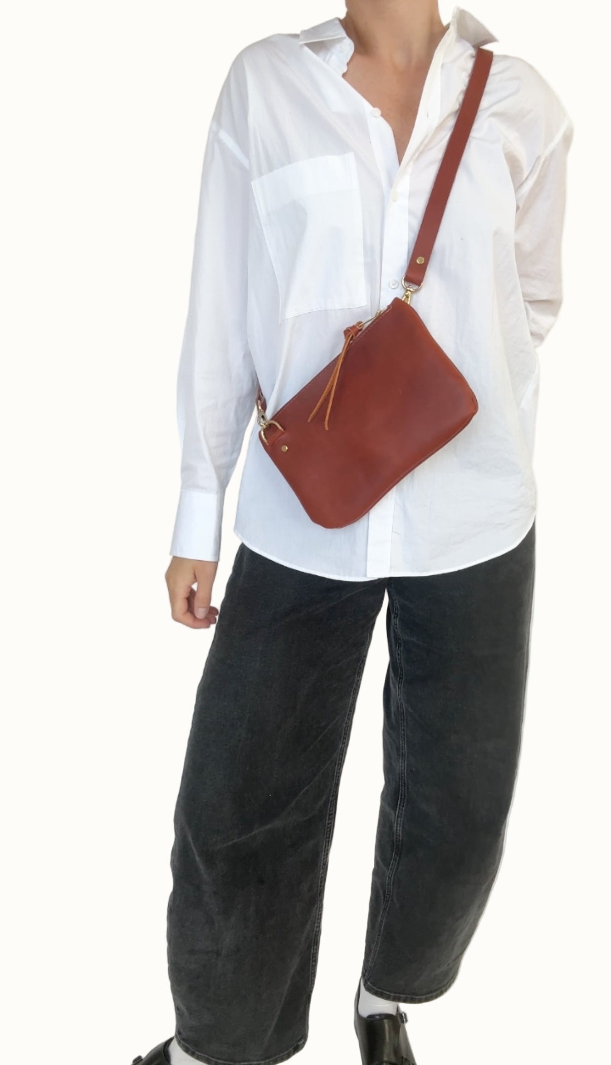 Person wearing a white shirt and dark pants with a brown leather bag on a plain background