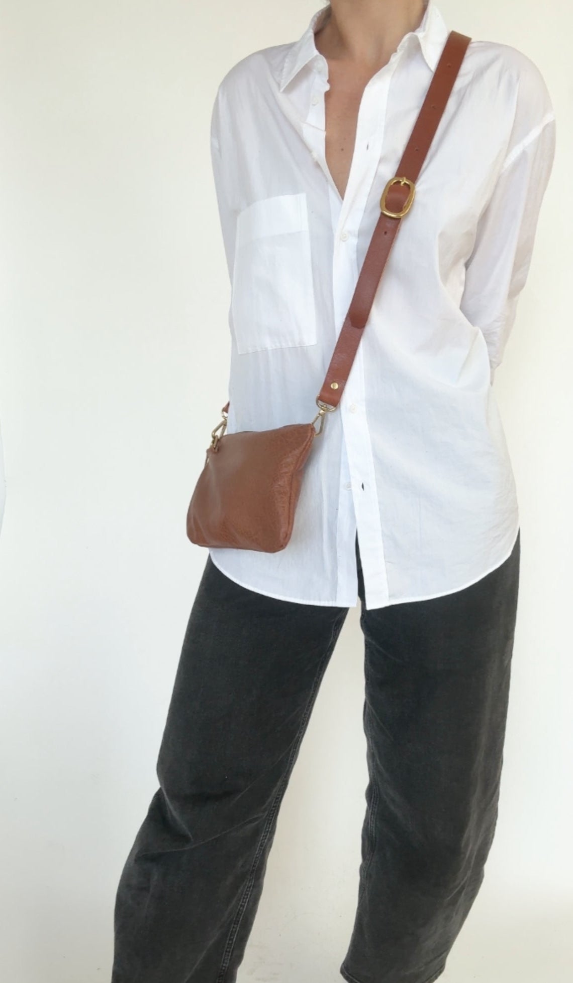 Person wearing a white shirt and black pants with a brown crossbody bag on a plain background