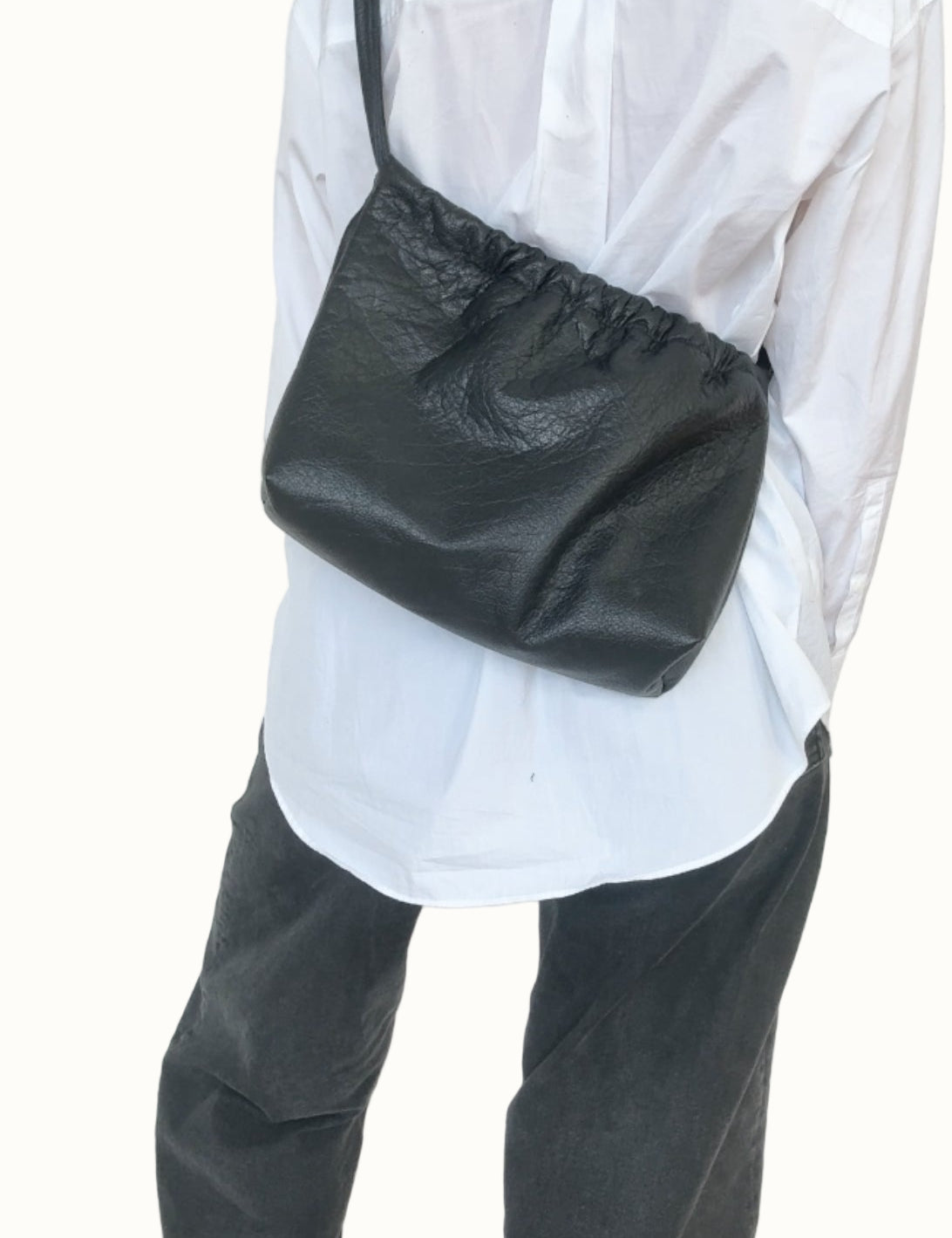 Person wearing a white shirt and dark pants with a black bag over one shoulder against a plain background