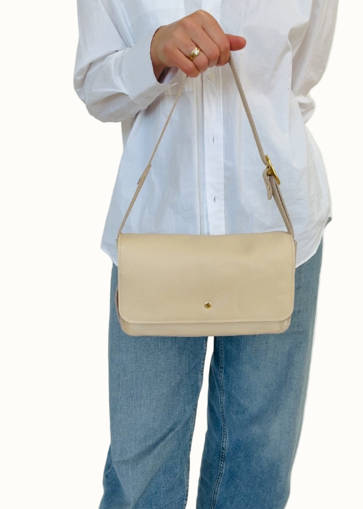A person wearing a white shirt and blue jeans is holding a cream-colored crossbody bag with a shoulder strap and a zipper closure.
