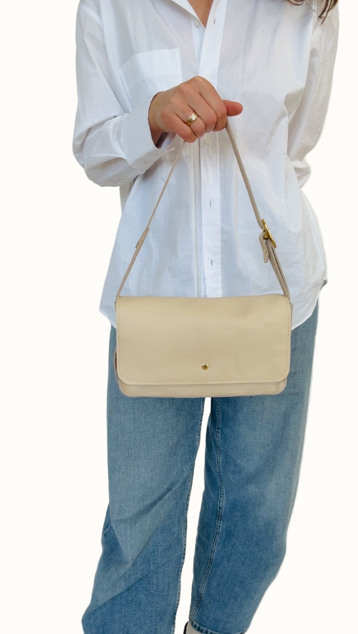 A person wearing a white shirt and blue jeans is holding a cream-colored crossbody bag with a shoulder strap and a zipper closure.